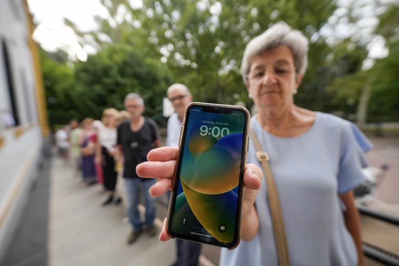 Una mujer hace cola a la espera de que abra sus puertas el colegio electoral de Ferroviarios de Córdoba y muestra la hora en su teléfono móvil momentos antes de entrar para ejercer su derecho al voto este domingo de elecciones generales en una jornada electoral marcada por el calor a lo largo del día en el que se espera que se alcancen los 40 grados