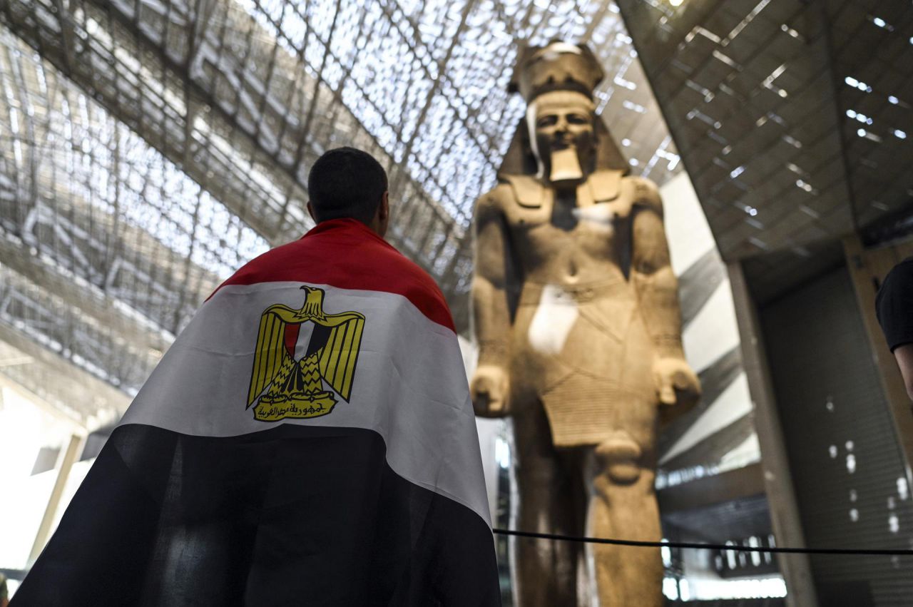Un visitante envuelto en una bandera observa la estatua de Ramsés II.