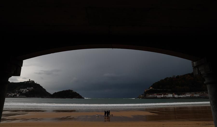 Varias personas caminan con sus perros por la playa de la Concha de San Sebastián, con los cielos cubiertos de fondo.